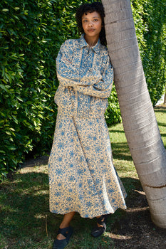 Woman leaning on tree wearing a blue and ecru tapestry jacket over a dress