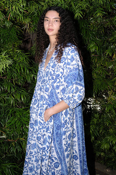 A woman in a garden with greenery wearing a caftan cut from a white with blue tapestry.