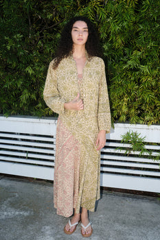 woman standing by white fence wearing a grren tapestry jacket over matching pink and green dress.