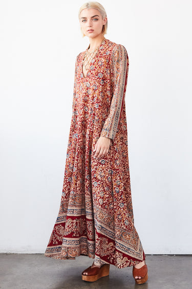 A woman in a studio with white walls wearing a red with blue flowers tapestry caftan with long sleeves.
