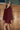 A woman wearing a burgundy long-sleeve mini dress with a textured, scalloped pattern, standing against a wooden fence background.