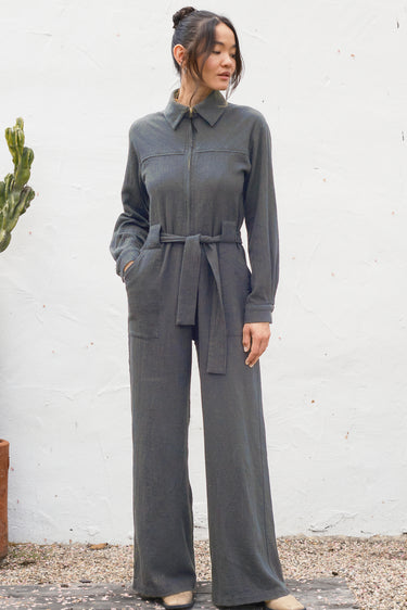Woman wearing a gray jumpsuit standing against a white wall.