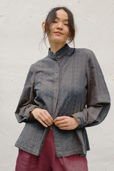 A woman wearing a dark gray patterned long-sleeve blouse with a high collar and button front, paired with burgundy pants, standing against a white textured wall.