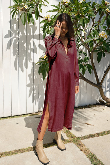 A woman wearing a long-sleeved, button-front burgundy maxi shirt dress with side slits, paired with beige ankle boots, standing outdoors in front of a white wooden fence and a leafy plant.