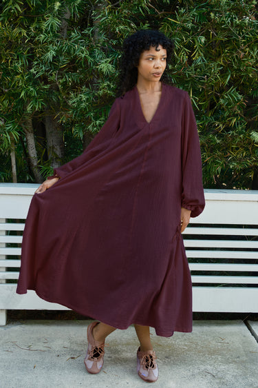 A woman wearing a loose, long-sleeved maxi dress in a black fig color, standing outdoors in front of a white bench and green foliage.