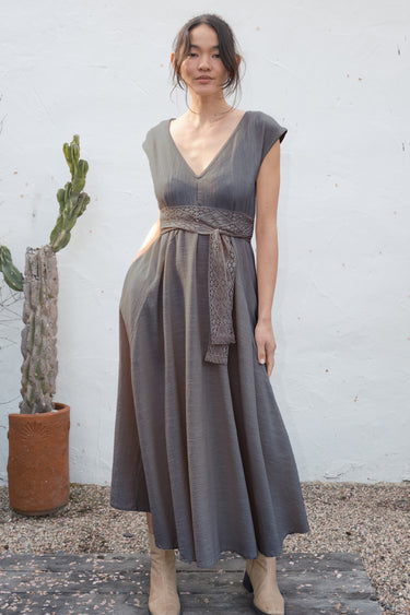 Woman wearing a sleeveless dress with a belt, standing against a white wall with a cactus plant.