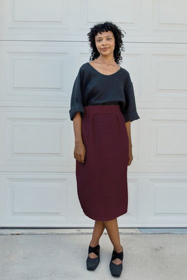 A woman stands in front of a white garage door wearing a black short-sleeve top and a high-waisted, knee-length burgundy skirt, paired with black Mary Jane shoes.