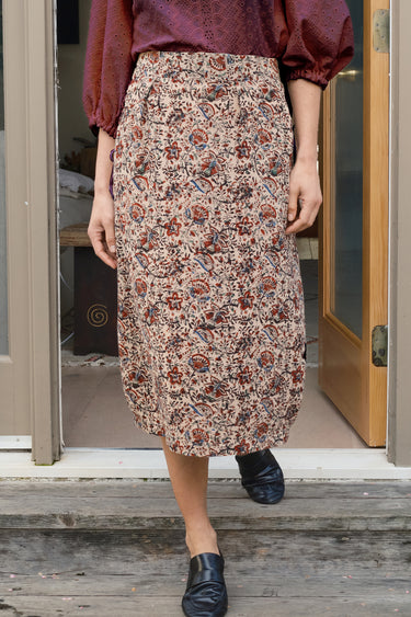 Person wearing a patterned skirt and maroon top standing on a wooden deck.