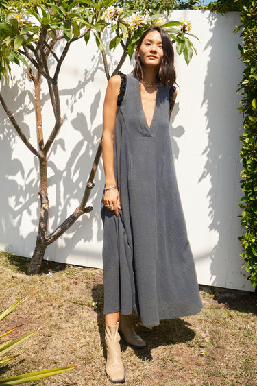 A woman wearing a sleeveless, deep V-neck midi dress in a textured gray fabric, standing outdoors against a white wall with green foliage.