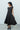 A woman wearing a black sleeveless midi dress with a boat neckline and a flared skirt, standing in front of a white garage door.