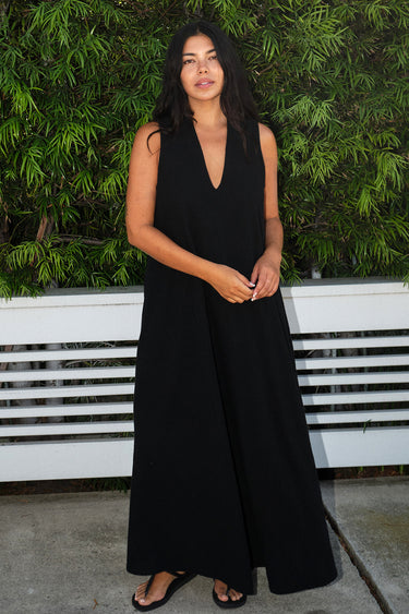 A woman wearing a black maxi caftan with deep cut arm holes and bias cut sides, featuring front cell phone pockets.