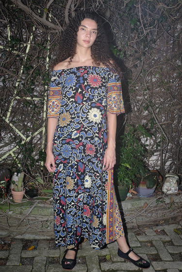 Woman standing in garden wearing a maxi dress with a bardot neckline, cut in a black ground sunflower tapestry.