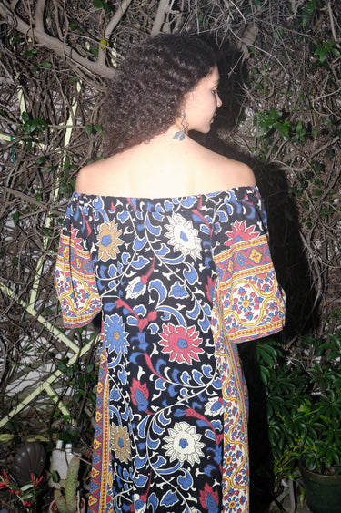 Woman standing in garden wearing a maxi dress with a bardot neckline, cut in a black ground sunflower tapestry. back view.