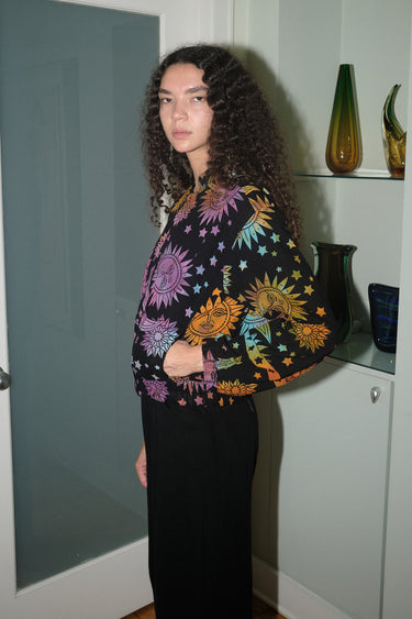A woman wearing a cropped black ground with rainbow suns, moons and stars tapestry jacket with a zip front, standing in a home environment.