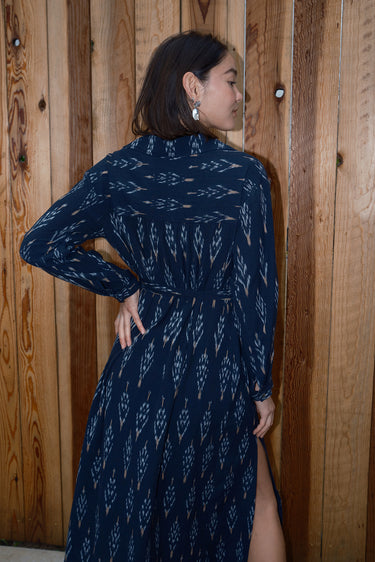 A woman standing in front of a wooden gate wearing black ikat shirt dress with long sleeves and an obi belt, back view.