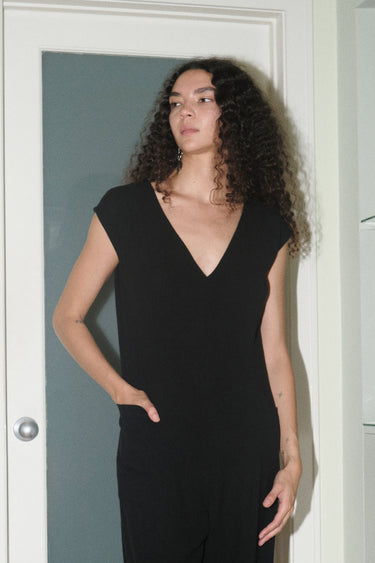 Close up of a woman standing in indoor setting wearing a slim black full length jumpsuit.