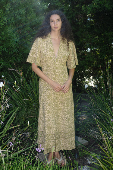 woman standing in garden wearing a light green print tapestry dress with zip front, v neck and elbow length sleeves