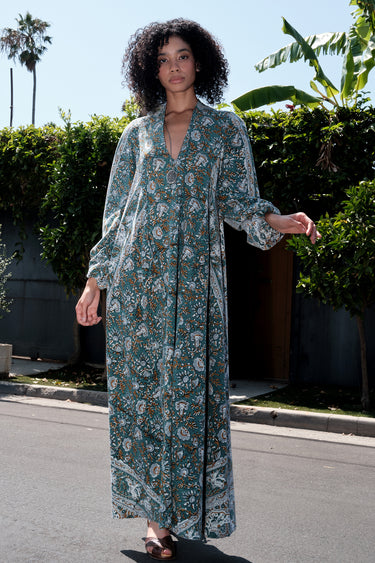 Woman wearing a long tarragon colored floral tapestry caftan standing outdoors with greenery in the background.