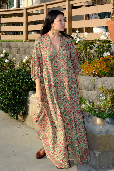 A woman standing outside wearing a beige ground with red and brown floral caftan.