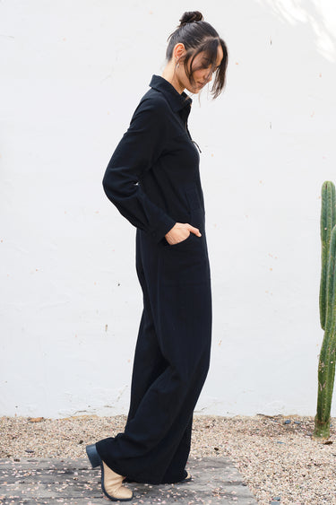 Person wearing a black jumpsuit standing against a white wall with a cactus plant.