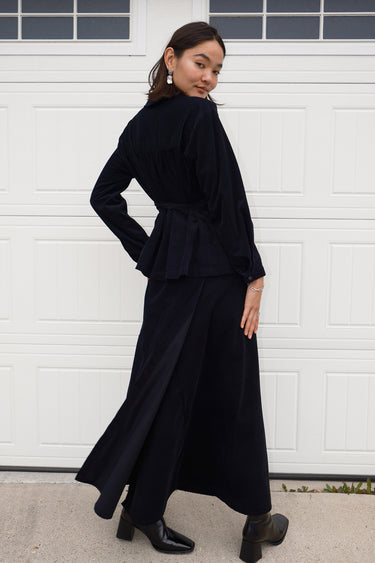 A woman standing in front of a white garage door, wearing a long, black, corduroy skirt with a V-neck top, three quester back view.