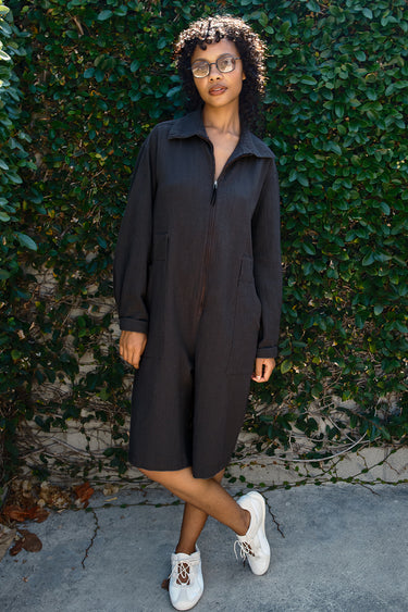 Person wearing a black jumpsuit standing against a green ivy-covered wall.