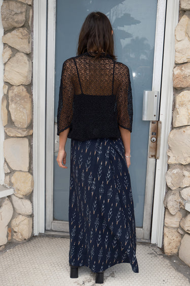 Woman standing a door way wearing a black zero waste lace shrug over a black tank top, paired with an ikat maxi skirt, back view.