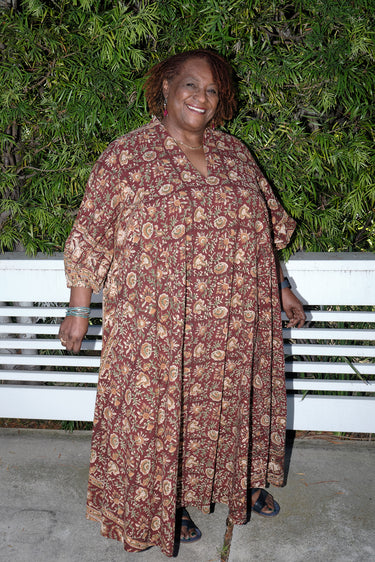 A woman outside standing against greenery wearing a raisin floral tapestry maxi caftan with three quarter balloon sleeves .