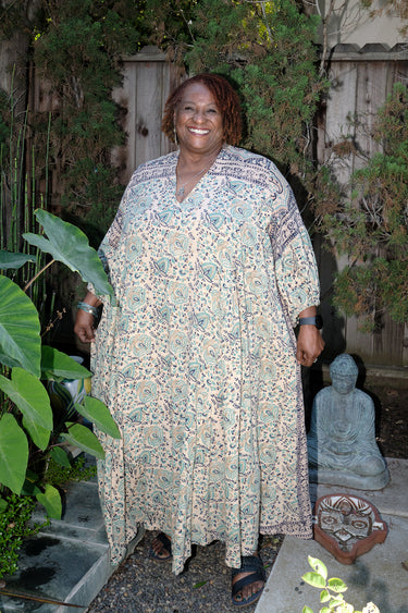 A woman wearing a maxi caftan with a floral pattern in a color that could be described as deep beige ground with tan, green and black paisley tapestry, standing outdoors.