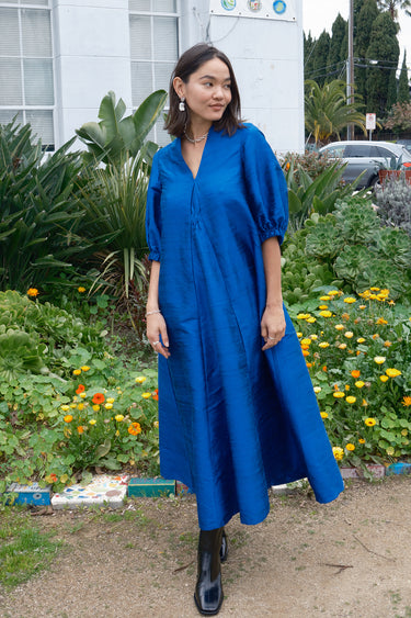A woman standing in a garden wearing a blue maxi dress with balloon sleeves and wide elastic cuffs, made of silk, posing next to a building with a blue sign on it.
