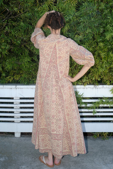 A woman standing outdoors wearing a blush maxi dress with floral hand block print design, featuring balloon sleeves and wide elastic at the cuffs, back view.