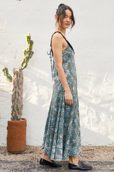 Woman wearing a floral dress standing next to a cactus plant against a textured white wall.