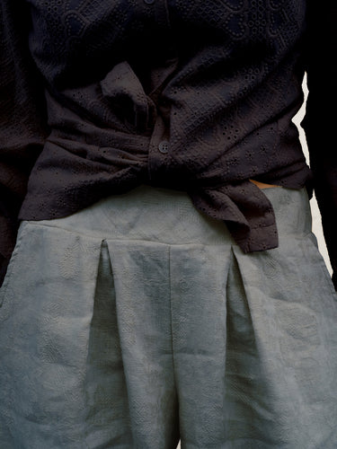 Close-up of a person wearing a dark charcoal textured button-up shirt tied at the waist and light gray pleated trousers.
