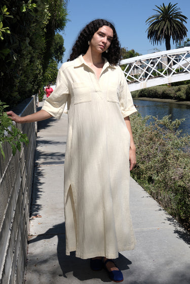 A woman standing on a pathway wearing an alabaster beige maxi shirtdress with a collar, sleeves, and a side slit, with a body made of crinkle textured osnaburg cotton.