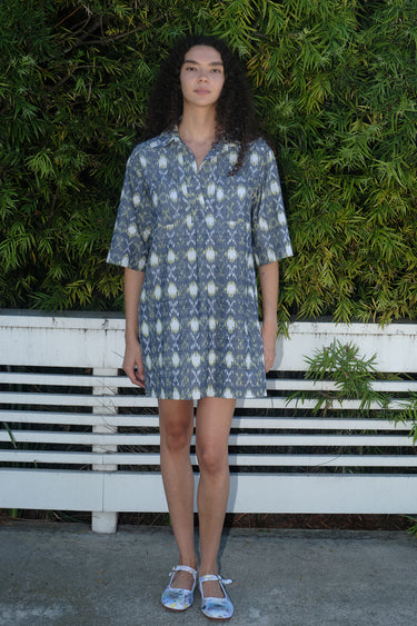 A person standing by a white fence with greenery wearing a mini shirt dress with a shirt collar and an "xo" Ikat pattern.