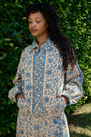 Woman wearing a blue and beige floral patterned outfit standing against a green leafy background