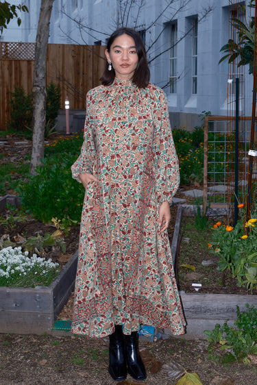Woman standing in garden wearing a long sleeve midi length dress in an ecru with red flower tapestry.