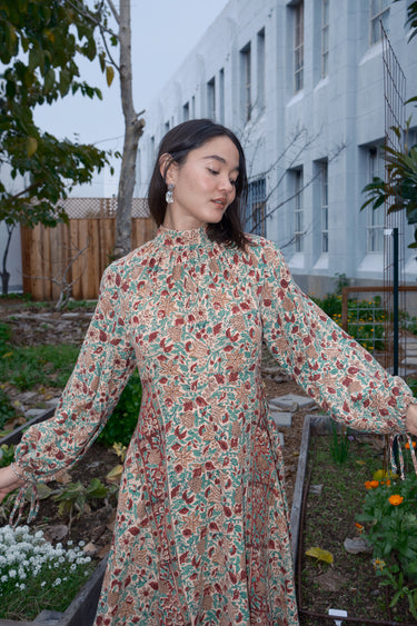 Woman standing in garden wearing a long sleeve midi length dress in an ecru with red flower tapestry.