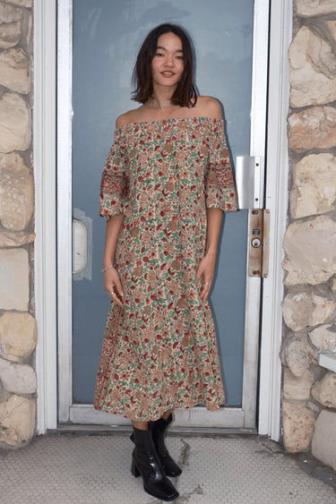 A woman standing in front of a door, wearing an off-the-shoulder floral tapestry dress with pockets, paired with boots.