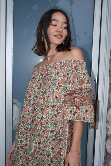 A woman standing in front of a door, wearing an off-the-shoulder floral tapestry dress.