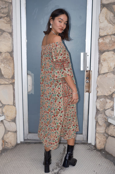 A woman standing in front of a door, wearing an off-the-shoulder floral tapestry dress with pockets, paired with boots, back view.