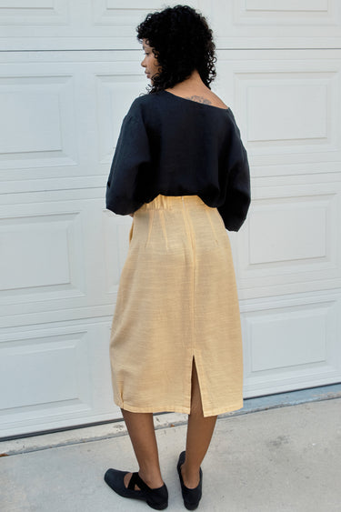 Person wearing a black top and yellow skirt standing against a white wall.