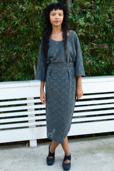 Woman wearing a gray lace tee and skirt standing in front of a white bench with green foliage in the background.
