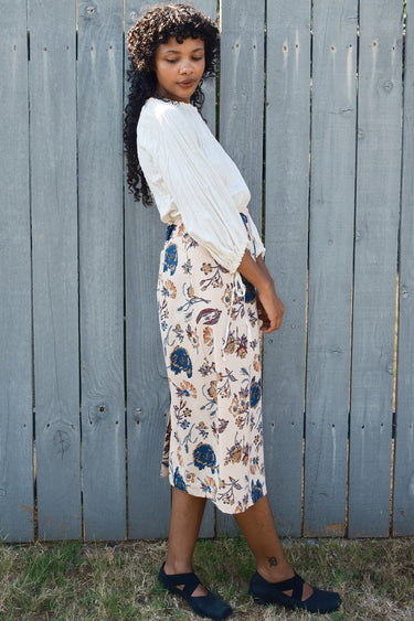 Woman wearing a white blouse and floral skirt standing against a wooden fence.