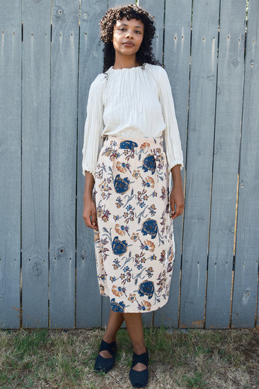 Woman wearing a floral skirt and white blouse against a wooden fence.