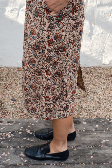 Person wearing a patterned skirt and black shoes on a wooden surface with a white background