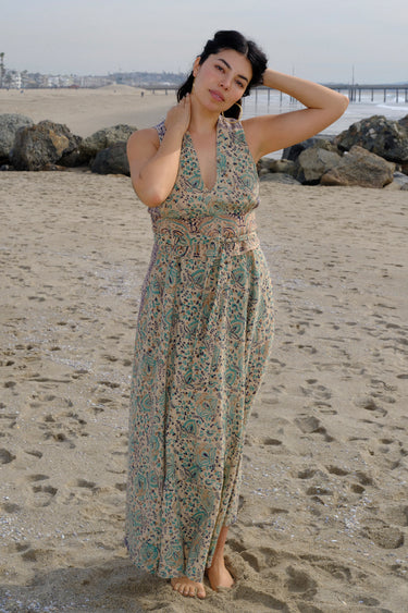 A woman at the beach wearing a sleeveless maxi caftan in a beige ground tapestry, turquoise flower handloom, featuring a matching obi belt.