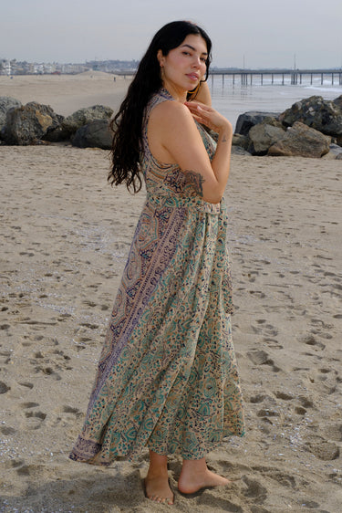 A woman at the beach wearing a sleeveless maxi caftan in a beige ground tapestry, turquoise flower handloom, featuring a matching obi belt. Side view