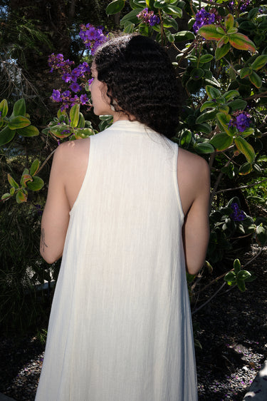 A woman outside wearing an alabaster off white maxi caftan with deep cut arm holes and bias cut sides, back view.