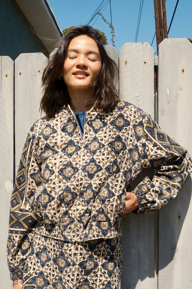A woman standing against a fence wearing a cropped, patterned tapestry jacket with a zip front and elastic sleeves.